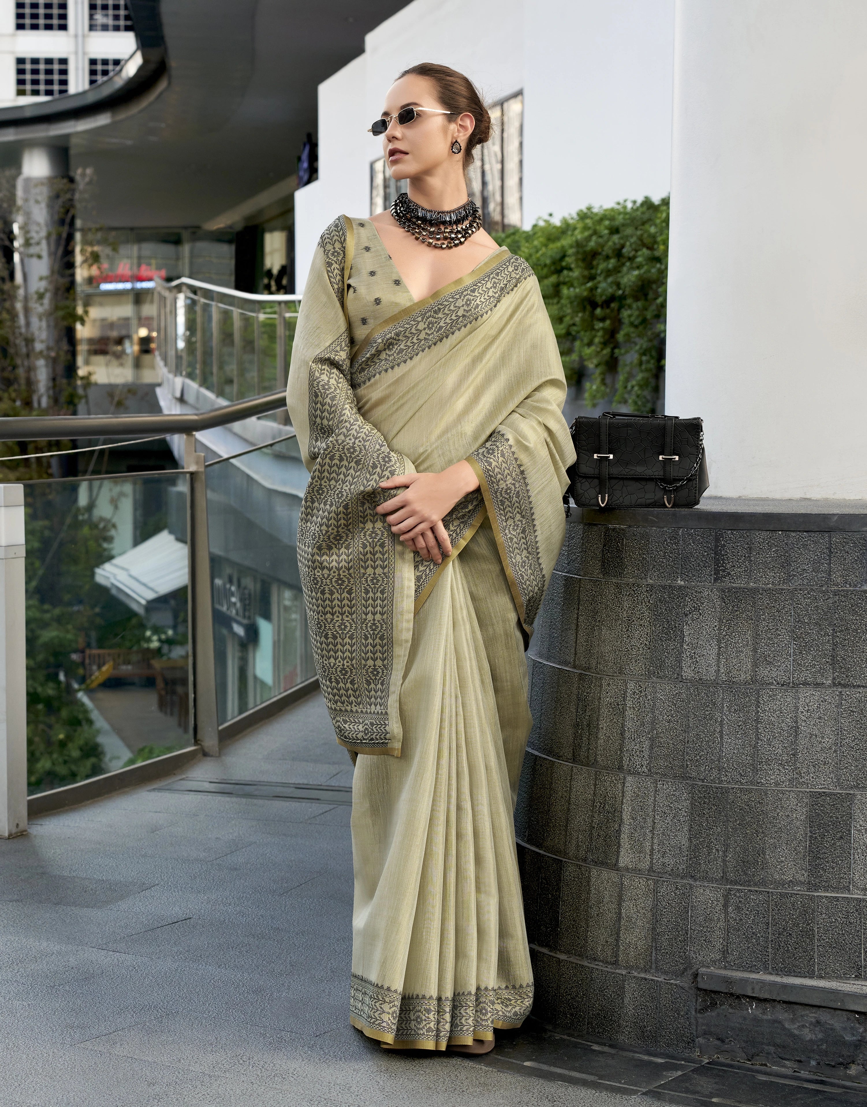 Woman in a light-colored saree standing outdoors near a modern building.